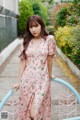 A woman in a pink floral dress leaning against a fence.