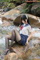 A woman in a school uniform sitting on a rock by a stream.