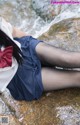 A woman in a school uniform sitting on a rock by the water.